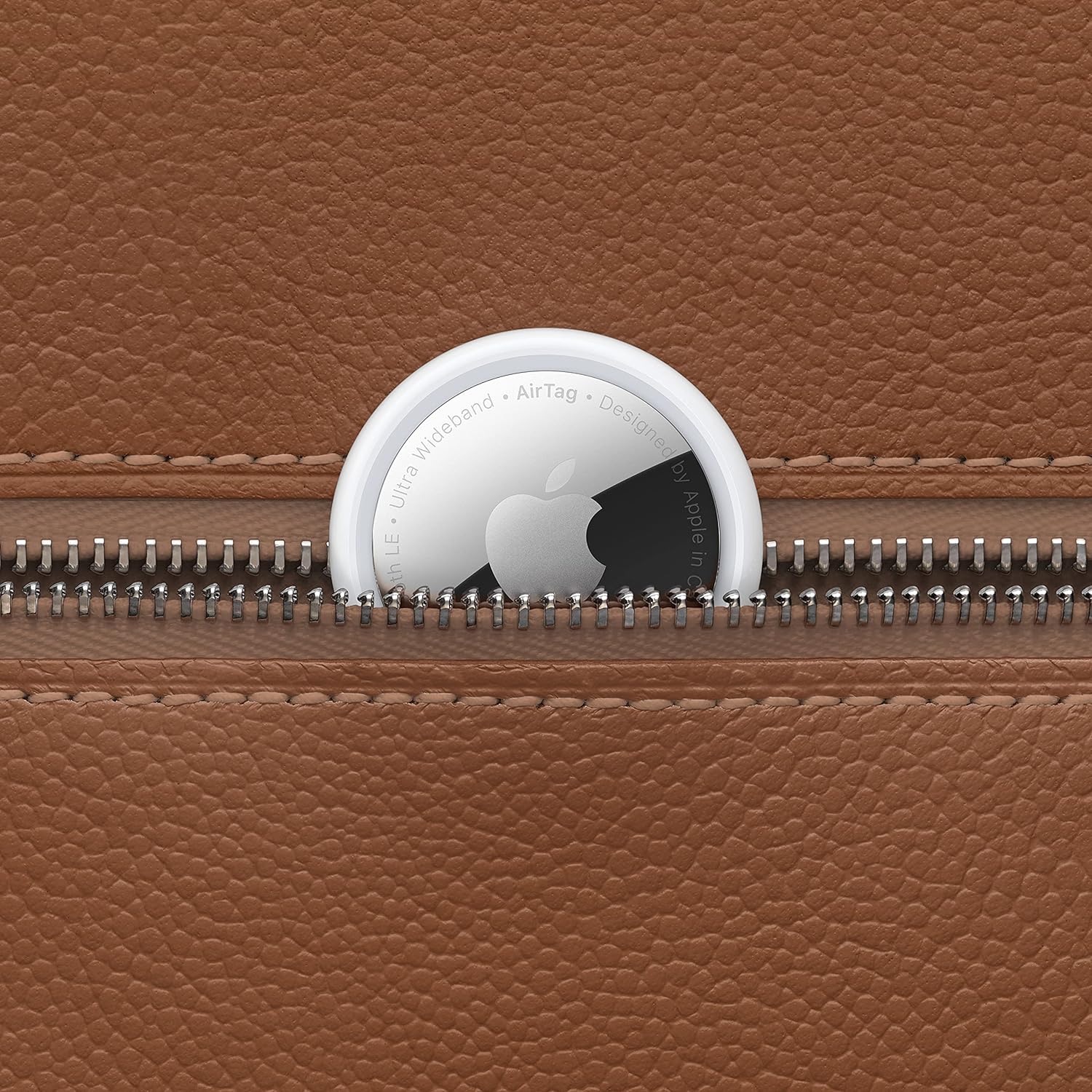 This image features an Apple AirTag, a small tracking device, placed over a textured brown leather surface with a metal zipper partly open. The AirTag is shown with its white side up, displaying the Apple logo, and text surrounding the logo providing information about the product, including mentions of 