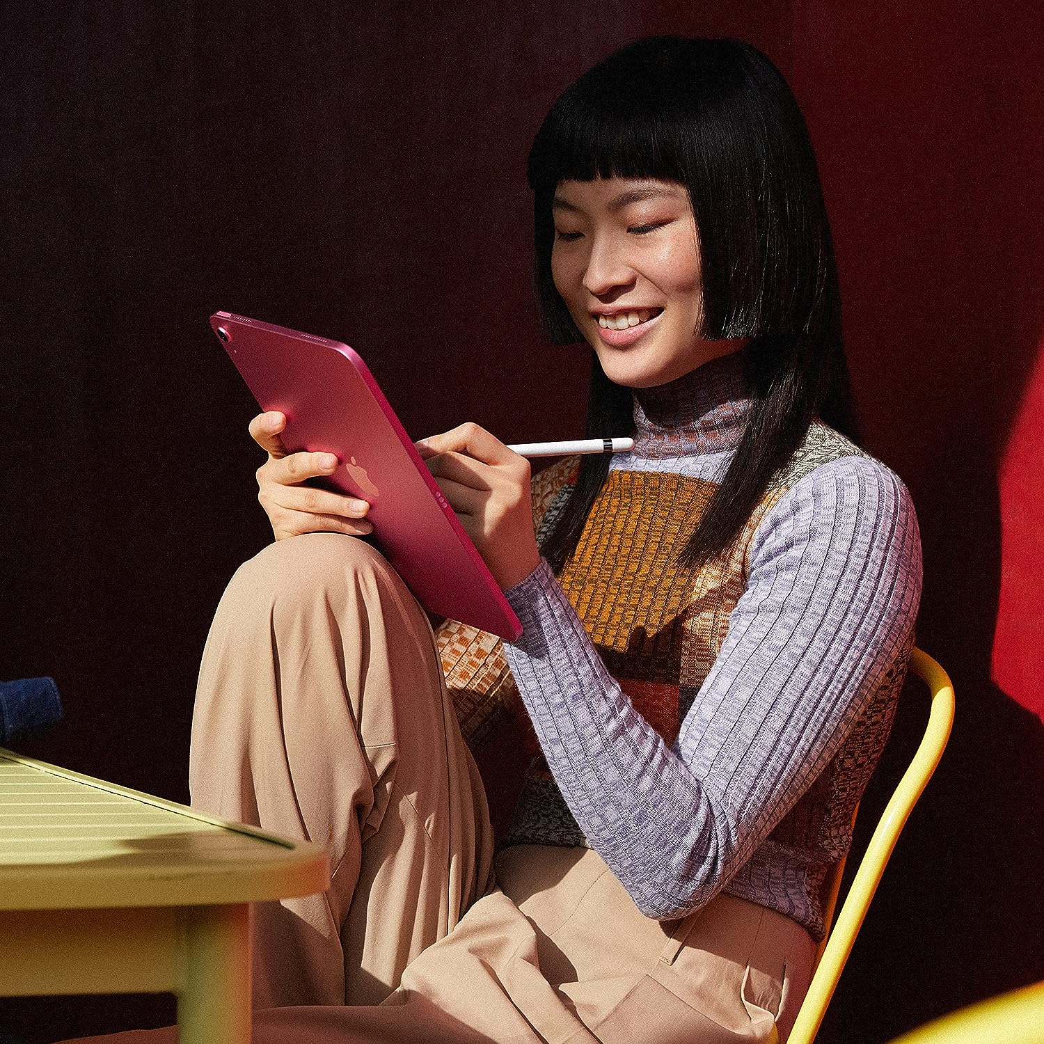 The image depicts a person sitting on a yellow chair, holding a pink iPad 10th Generation with a stylus. They appear to be smiling and engaged with the content on the screen. The individual has black hair with a straight fringe, is wearing a multicolored top layered with a lilac turtleneck, and beige trousers. The background consists of red and maroon tones, providing a contrast that highlights the subject. The scene suggests a comfortable and relaxed setting, possibly indicating a casual work environment or a personal leisure activity.