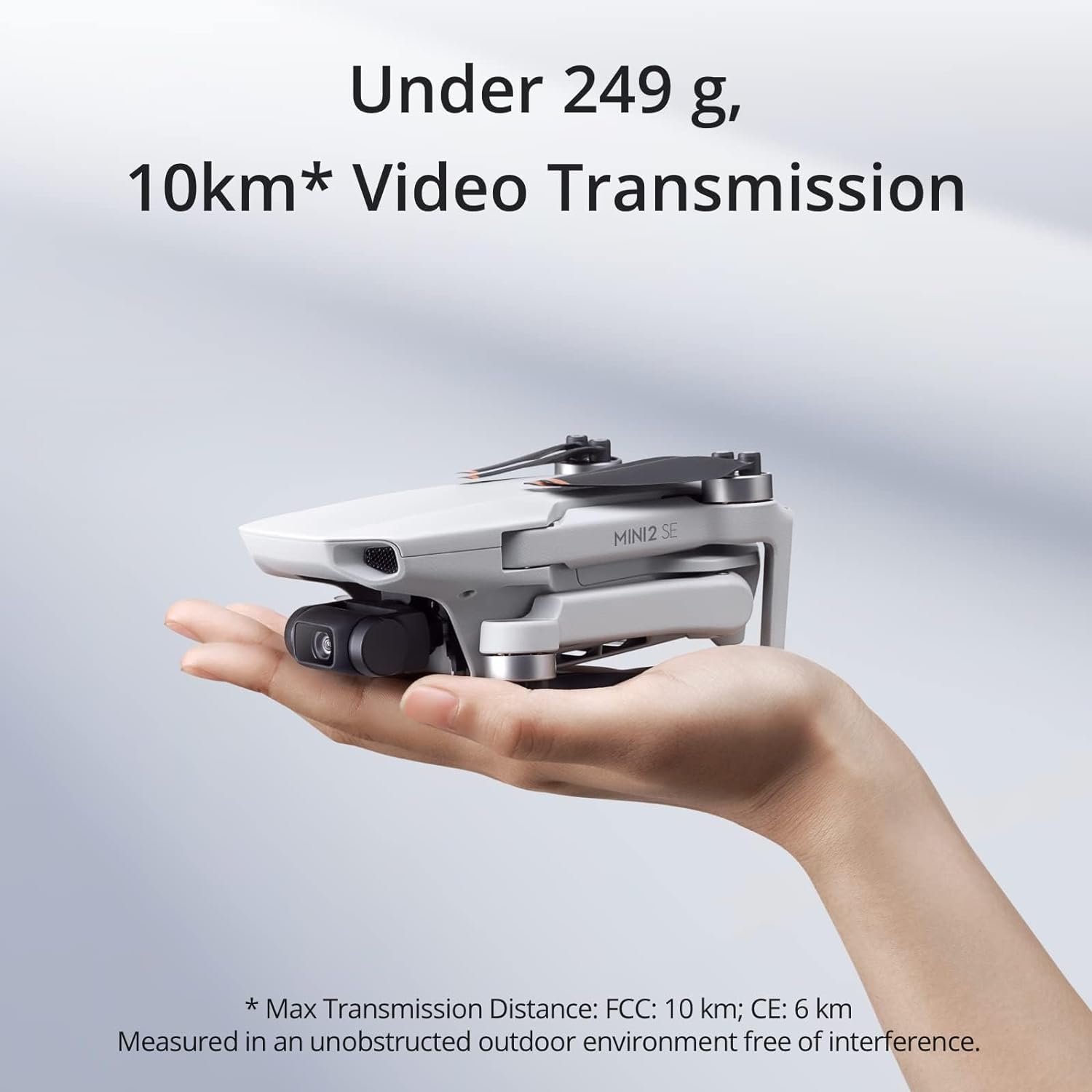 This image features a hand holding a DJI Mini 2SE drone, specifically labeled as the MINI 2 SE. The advertisement highlights that the drone weighs under 249 grams and boasts a 10-kilometer video transmission capability, with a note that the maximum transmission distance is subject to different regulations (FCC: 10 km; CE: 6 km) and is measured in an unobstructed outdoor environment free of interference. The background is a soft, nondescript gradient, focusing attention on the drone and its features.