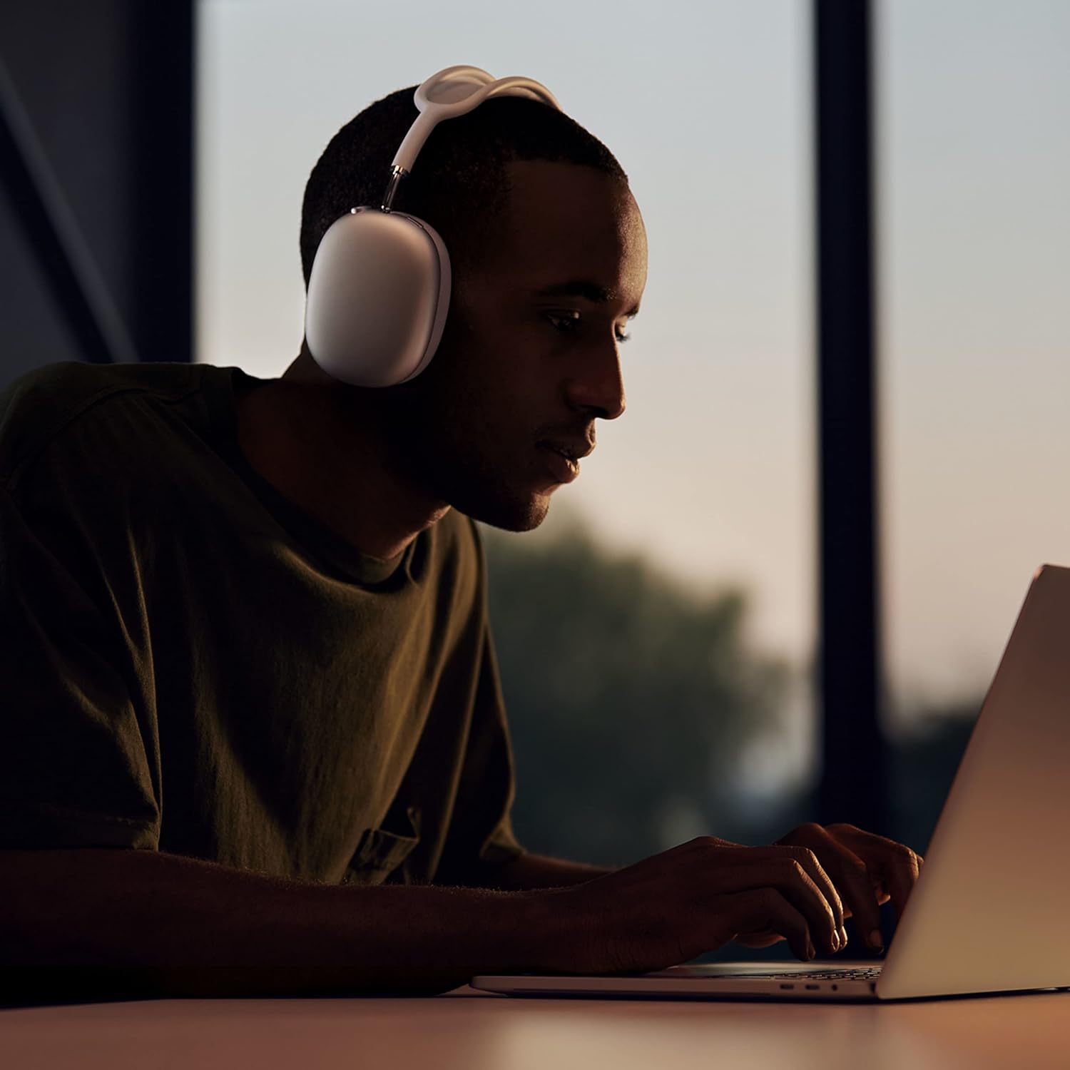 This image shows a person wearing Apple AirPods Max, engaged in using a laptop. The setting appears to be indoors with natural lighting coming from the window, suggesting it could be daytime. The person has a focused expression, indicating concentration on the task at hand on the laptop. The exact details of what's on the laptop screen are not visible in this image.