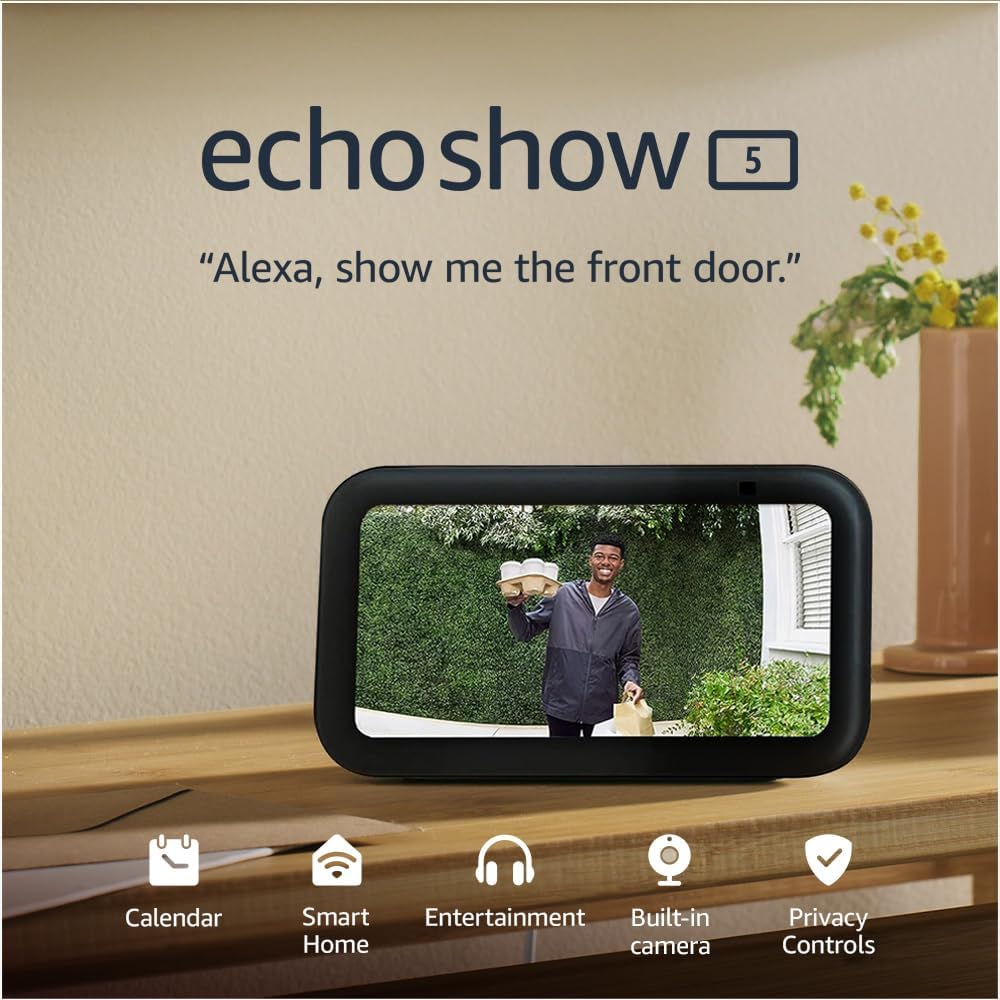 This is a promotional image for the Echo Show 5, a smart display that integrates Amazon's virtual assistant, Alexa. The device is shown on a wooden surface displaying a live camera feed of a man standing at a front door, holding a package and a bag, seemingly making a delivery. The screen of the Echo Show 5 also indicates some features such as Calendar, Smart Home, Entertainment, Built-in camera, and Privacy Controls. There's also a graphic in the bottom right corner indicating the enhancement of privacy controls. The text in the image suggests that the device can be used to view a camera feed by giving a voice command to Alexa.