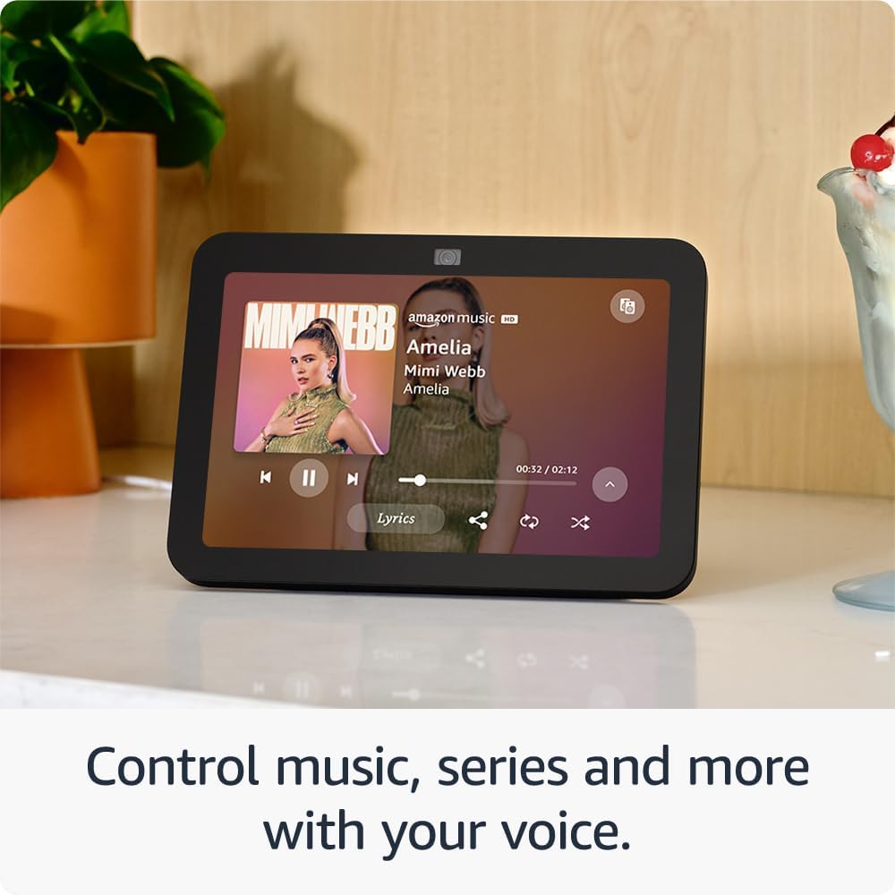 The image displays a smart display on a beige tabletop against a backdrop featuring a plant in a terracotta pot and a decorative bird figurine. The smart display screen shows a music application interface with an album cover featuring a female artist, along with playback controls and the track title “Amelia” by “Mimi Webb.” There is a caption at the bottom of the image that reads, “Control music, series and more with your voice,” suggesting that the smart display is equipped with voice control capabilities, likely for various multimedia content.