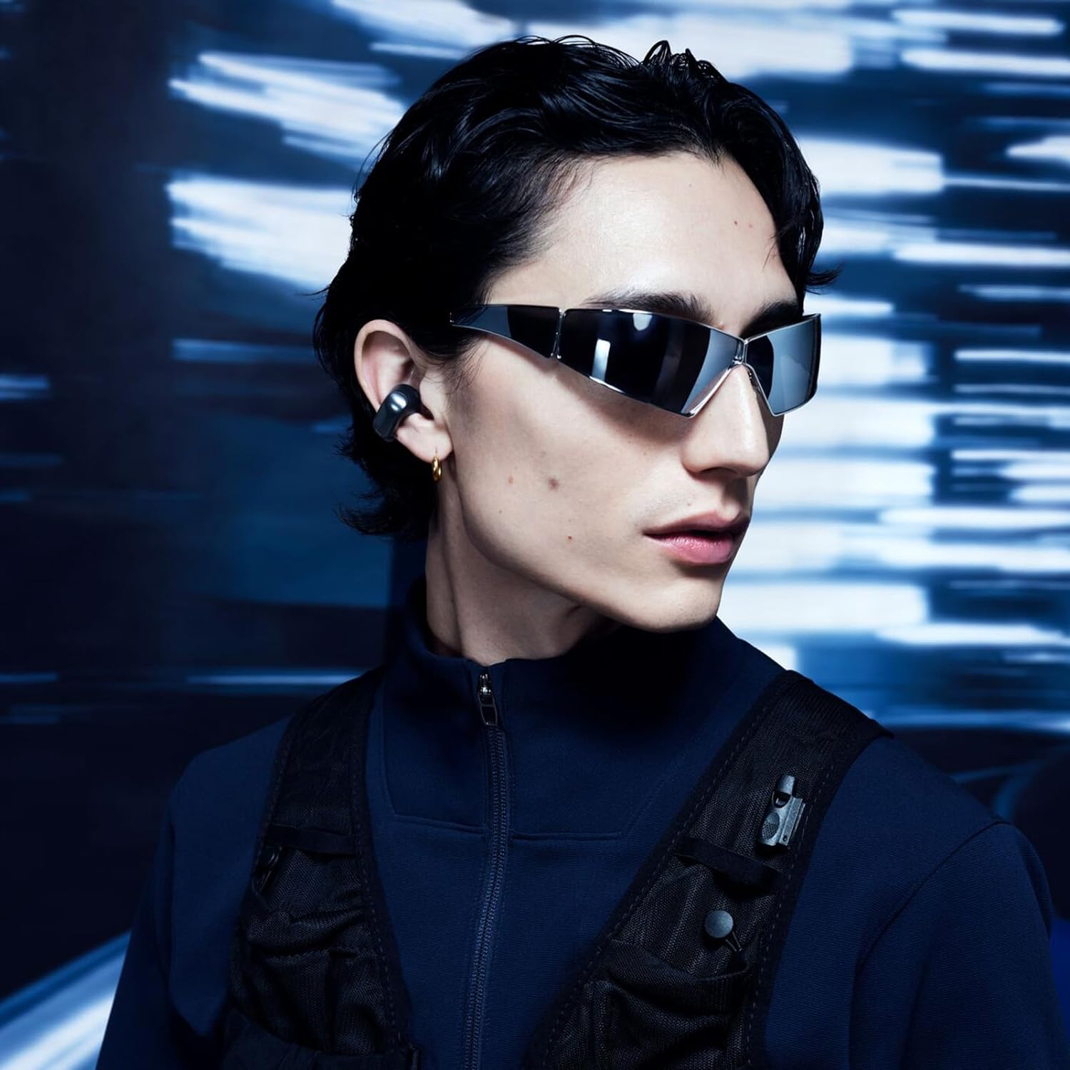 This image shows a person with a slicked-back hairstyle wearing futuristic-looking sunglasses and what appears to be wireless earbuds. The individual is also dressed in a navy blue high-neck jacket with what looks like a harness or detailed vest over it, featuring some pockets and metallic attachments. The background suggests motion, with light streaks giving a sense of speed or digital data flow. The overall image exudes a modern, tech-inspired vibe.