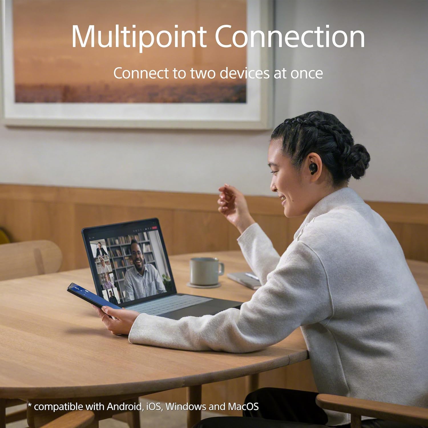 The image depicts an indoor setting where a woman is sitting at a wooden table, engaging in a video call with a man on her laptop. She is also holding a smartphone in her hand, which seems to be connected to the call as well. The woman is wearing a white jacket and has her hair styled in a braid. She wears earbuds, suggesting she might be using a multipoint connection feature that allows her to connect to both the laptop and the smartphone simultaneously.

On the table, there is a grey cup, suggesting she could be enjoying a beverage during the call. In the background, there's a framed artwork on the wall, and the room appears to be well-lit with natural light. The text 
