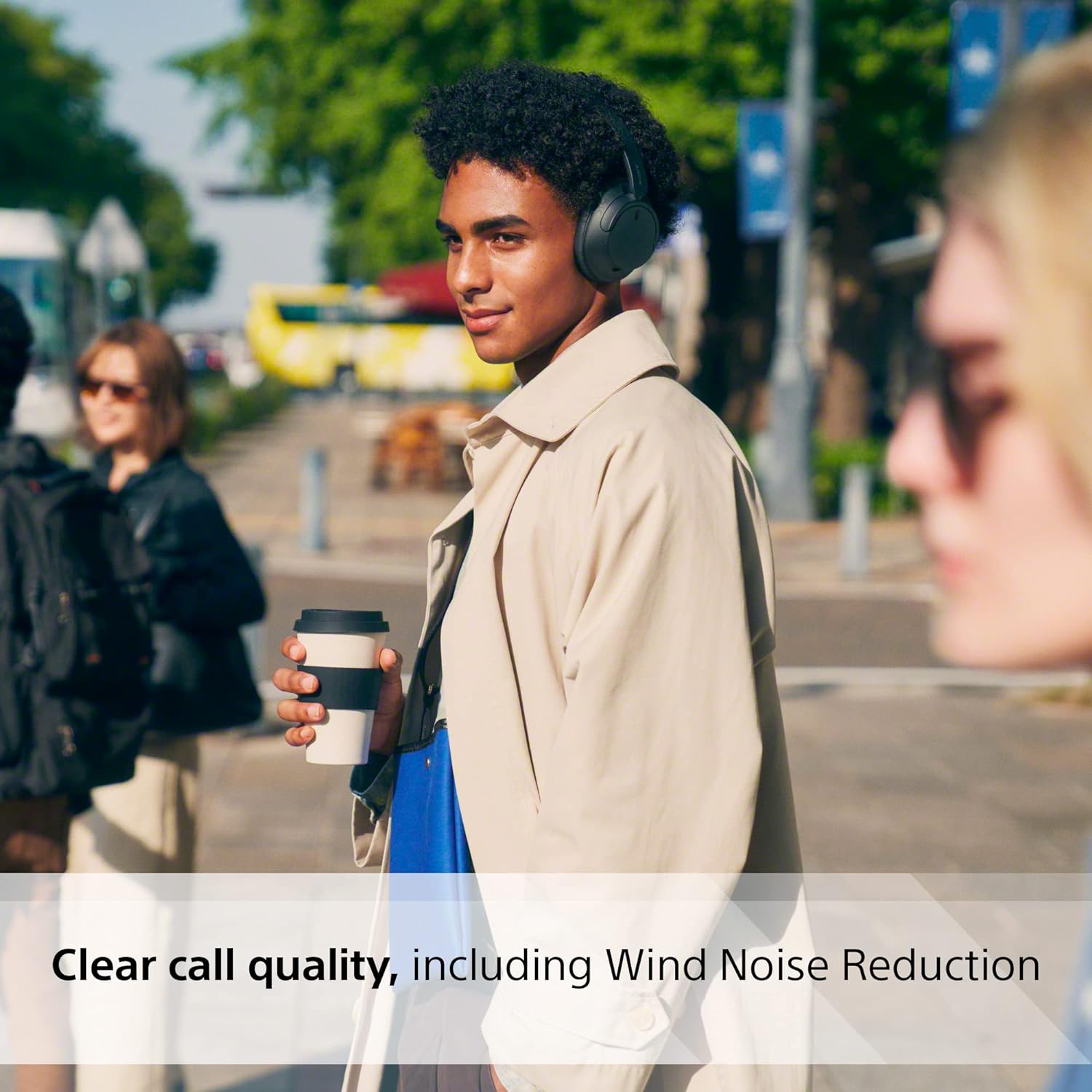 In this image, there is a young person walking outdoors on a sunny day, wearing a pair of over-ear Sony WH-CH720N headphones. They are holding a takeaway coffee cup in one hand and appear to be enjoying their walk, with a slight smile on their face. The person is stylishly dressed in a light beige or tan trench coat over a blue garment. In the background, there are other people who seem to be going about their day, with one person partially out of focus in the foreground.