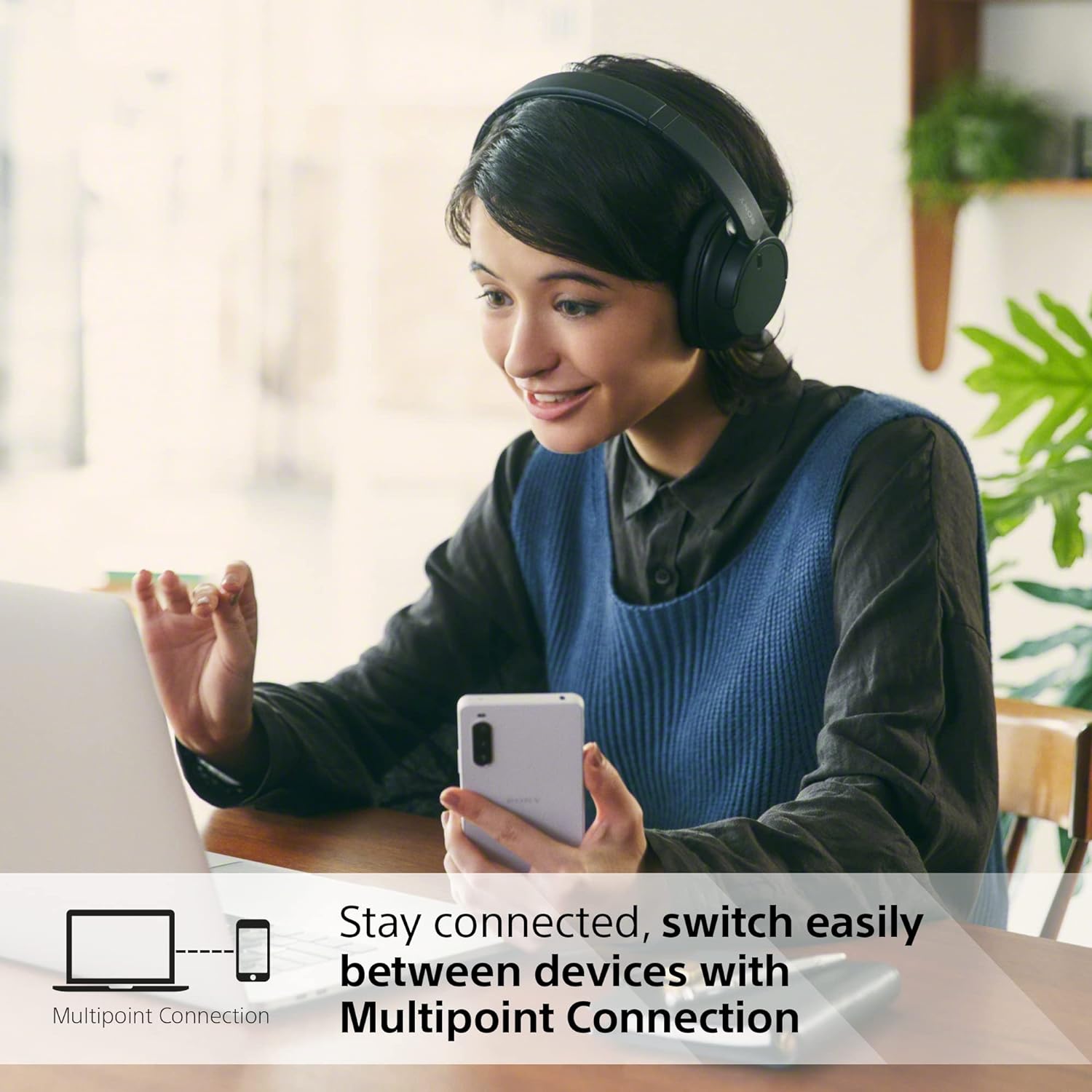 The image shows a woman sitting at a desk wearing a pair of over-ear Sony WH-CH720N headphones. She is holding a smartphone in one hand and gesturing with the other, appearing to be engaged in a conversation or making a video call. The woman is smiling and looking at what might be a computer screen, out of the frame. There are plants and a blurred background suggesting an office or a home environment. On the bottom left, there's an icon that implies connectivity between a computer and a smartphone.