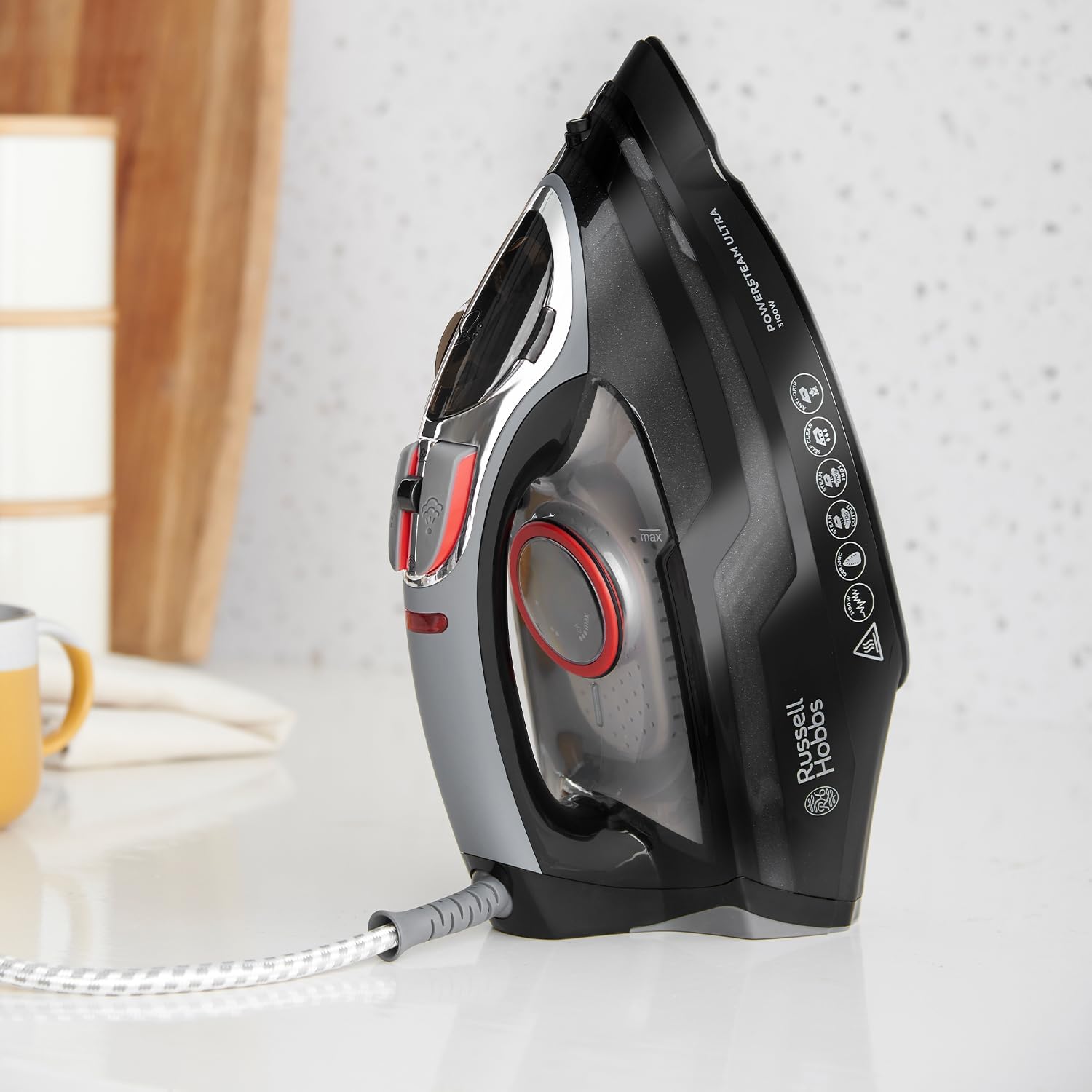 This image shows an electric steam iron sitting on a kitchen countertop. The iron is predominantly black with red accents and has a shiny finish, indicating that it's likely made of a high-grade plastic with some metallic components. Various control settings and labels can be seen on the iron, including steam and temperature controls. It has a power cord that extends from the back, and it appears to be a modern appliance, designed for ironing clothes to remove wrinkles. The brand 