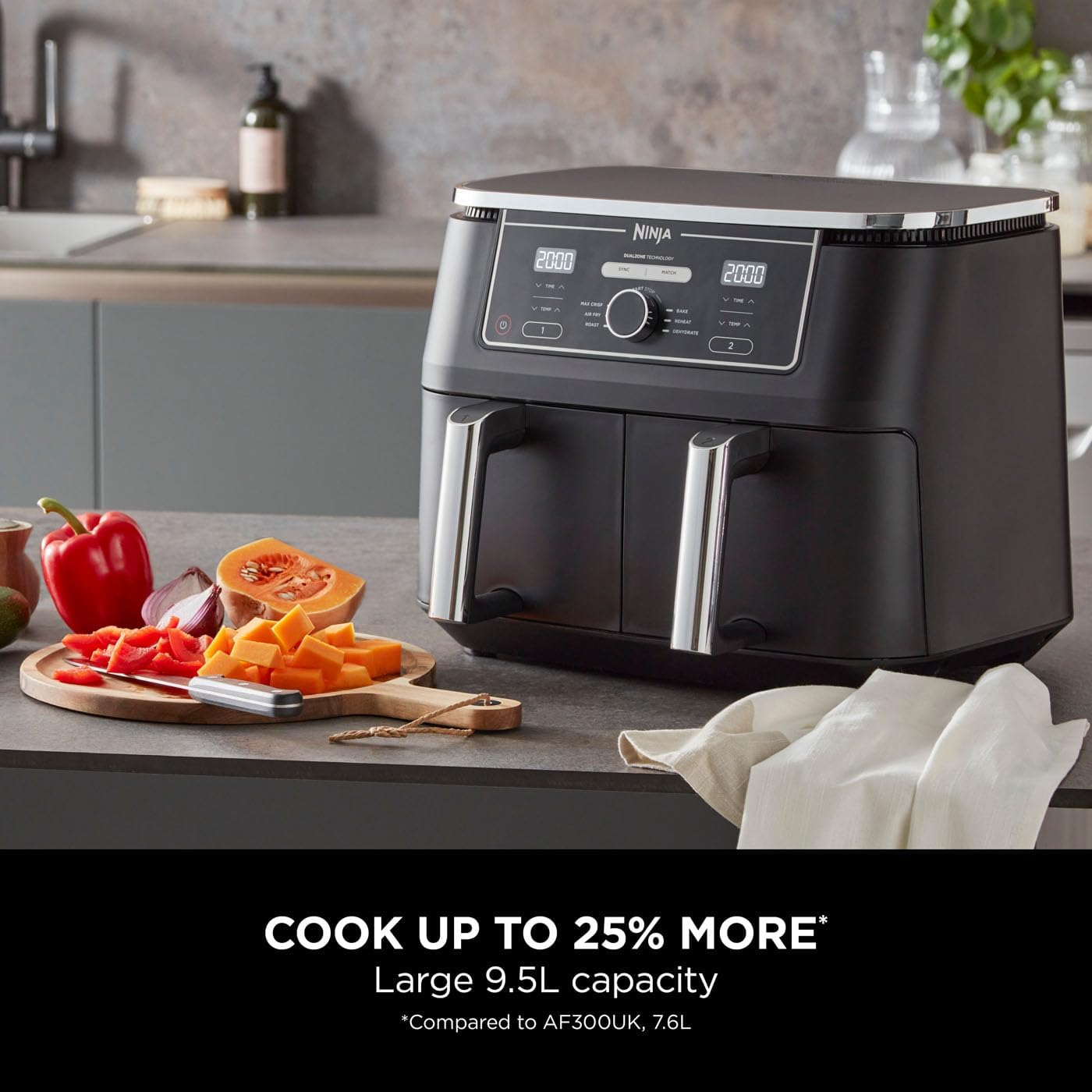 The image shows a modern kitchen countertop with a Ninja Dual Air Fryer 9.5 L. The air fryer is black with digital controls at the top, and it appears to have dual baskets with stainless steel handles. To the left of the air fryer, there's a cutting board with sliced red bell pepper, a piece of papaya, a piece of butternut squash, and a red apple, along with a knife placed over the board. A white kitchen towel is also partially visible in front of the countertop, below the air fryer.