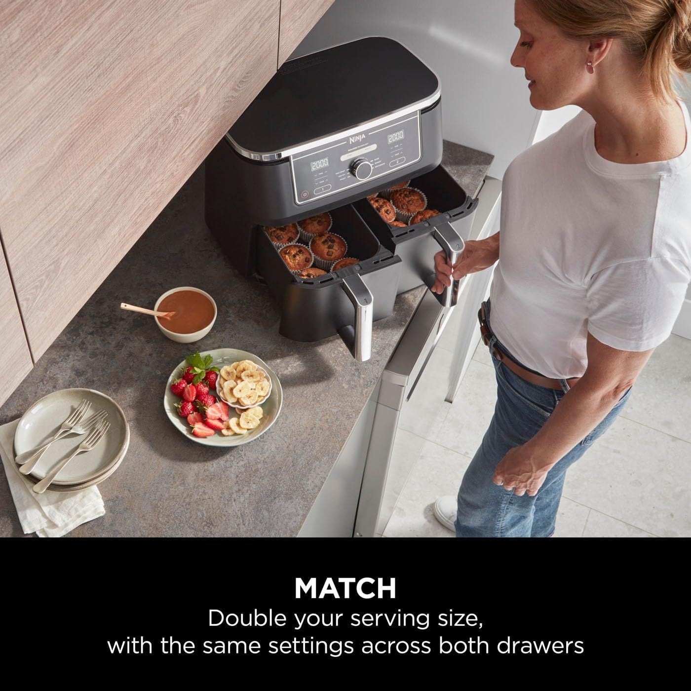The image shows a kitchen counter with a Ninja Dual Air Fryer 9.5 L. There are food items being cooked in both baskets, which appear to be bagels or some form of baked goods with a hole in the center. A woman is standing by the appliance, seemingly interacting with it or checking the food. On the countertop, there is a plate with strawberries and banana slices, a bowl possibly containing some type of sauce or dip, and a set of dining utensils on a napkin.
